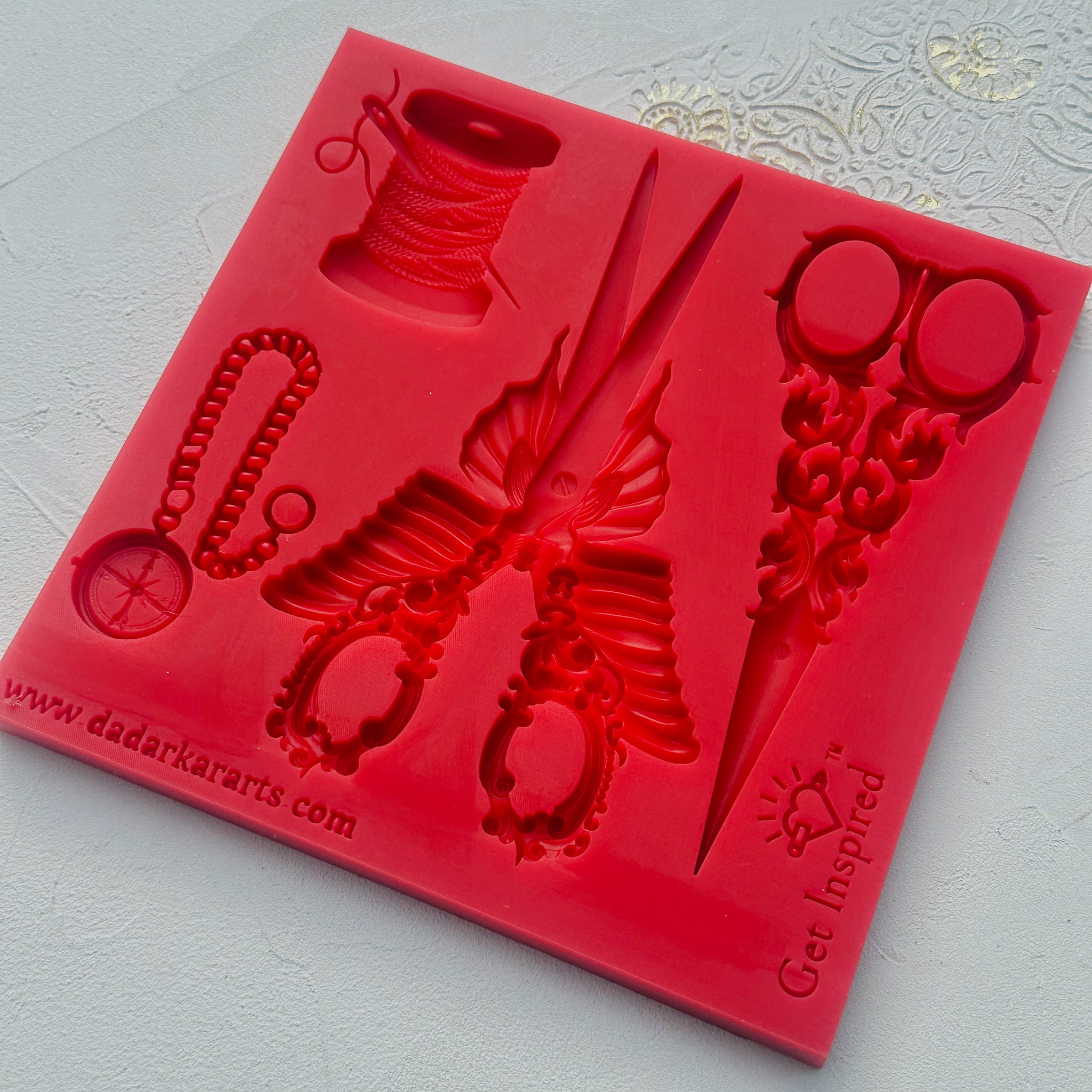 A red silicone mold featuring 2 steampunk style sewing scissors, a spool of thread with a needle in it, and a small pocket compass is against a white background.