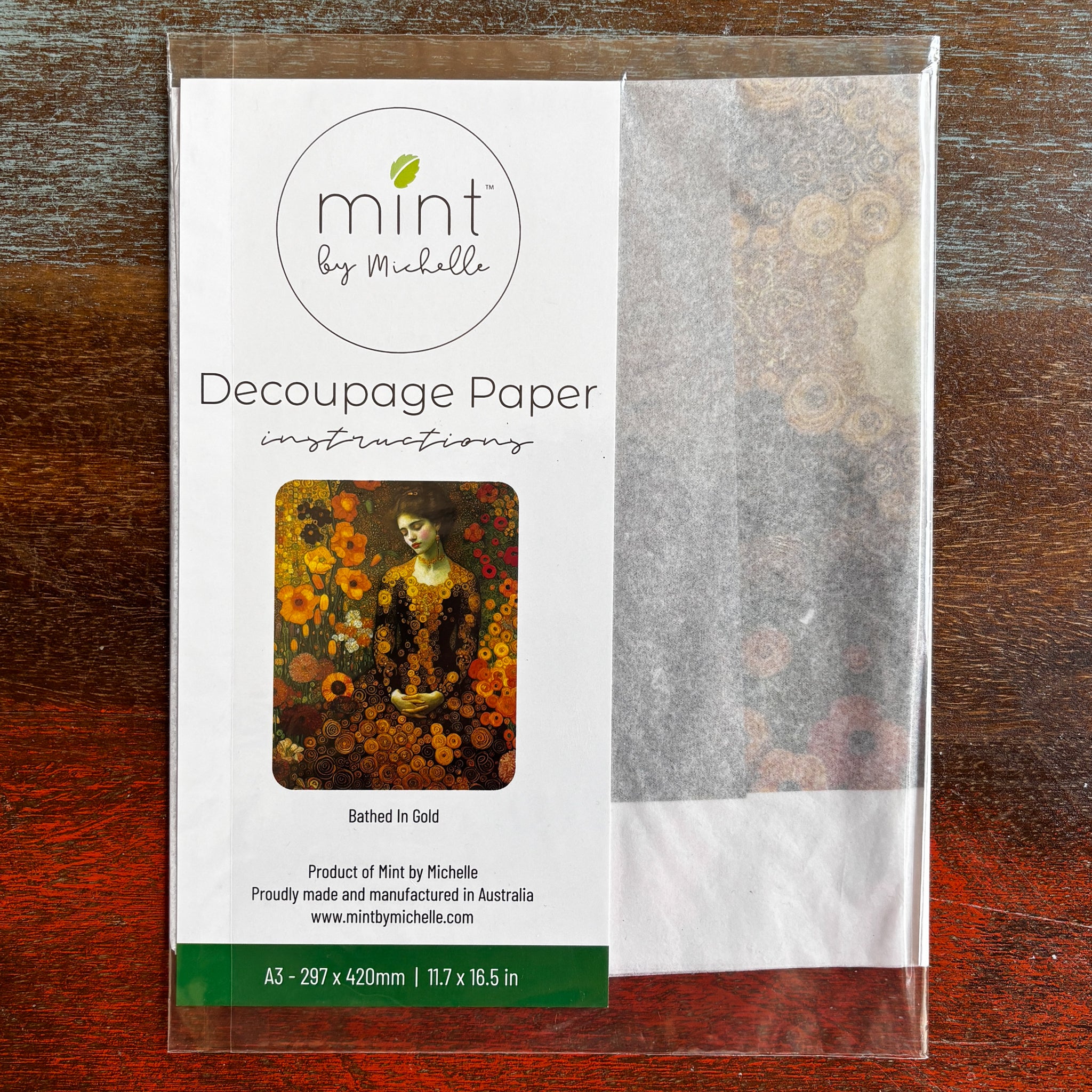 A package of Mint by Michelle's Bathed in Gold A3 decoupage paper is against a dark wood background.