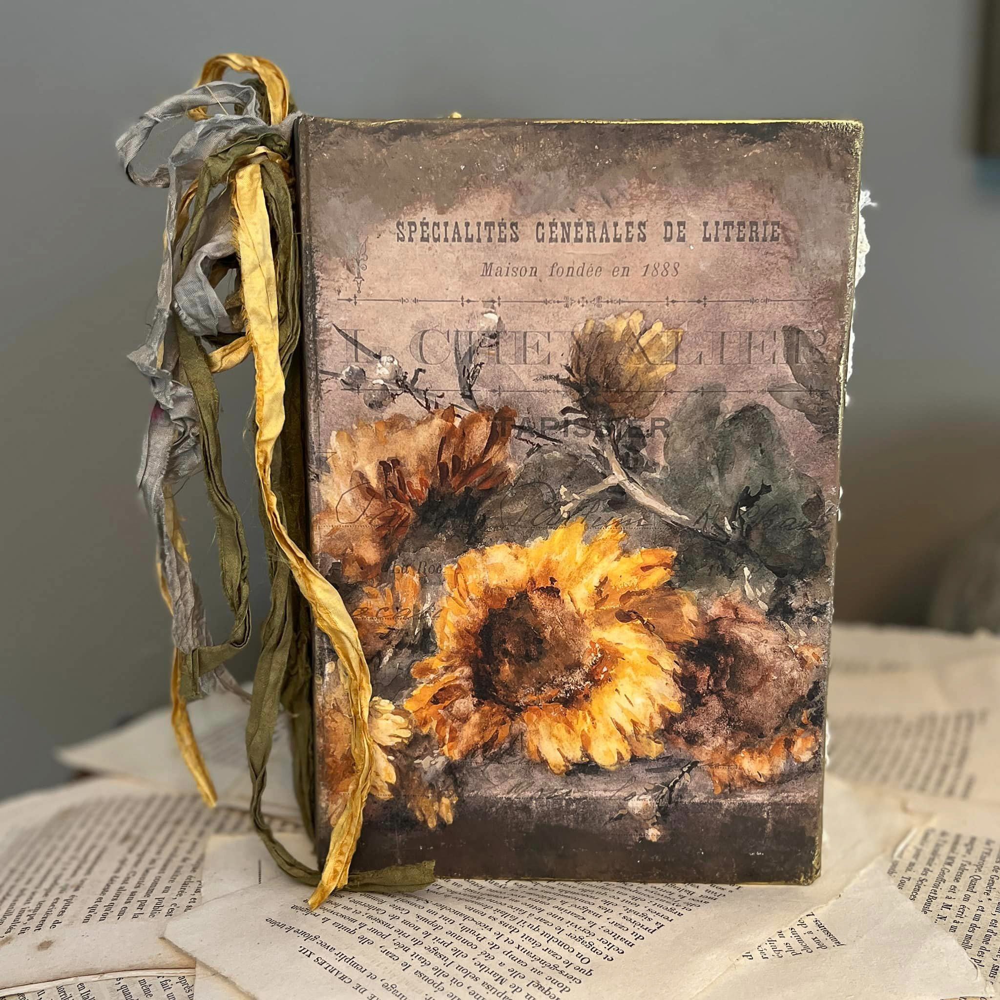 A vintage book features Decoupage Queen's Rochelle rice paper on its cover. Tattered ribbon is at the top of the book binding.