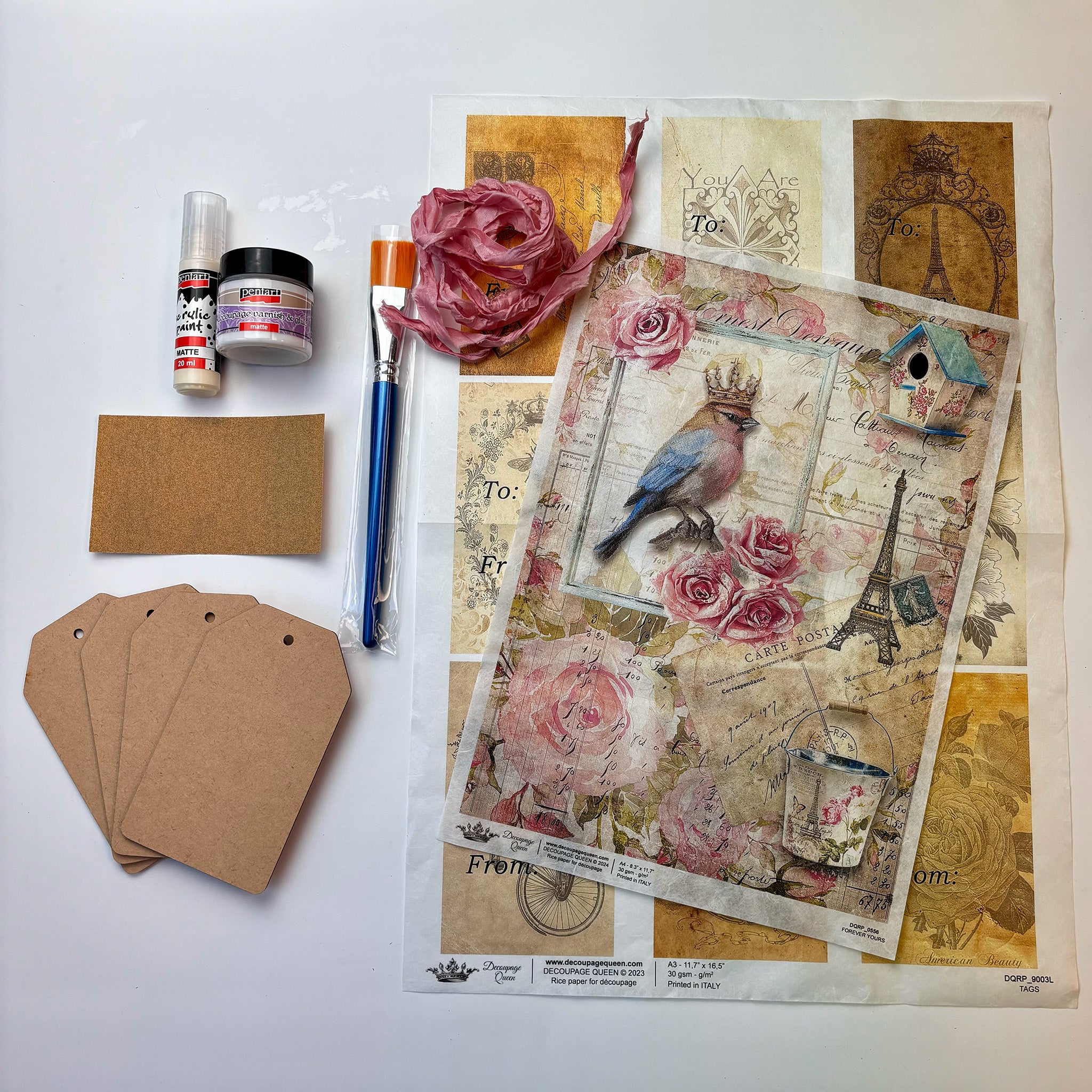 Against a white background is a decoupage beginner's kit that includes 4 wood gift tags, a sheet of To and From rice paper tags, decoupage glue, acrylic paint, a paintbrush, and an A4 rice paper featuring a French inspired design that includes pink roses, the Eiffel Tower, a birdhouse, and a bird wearing a crown.