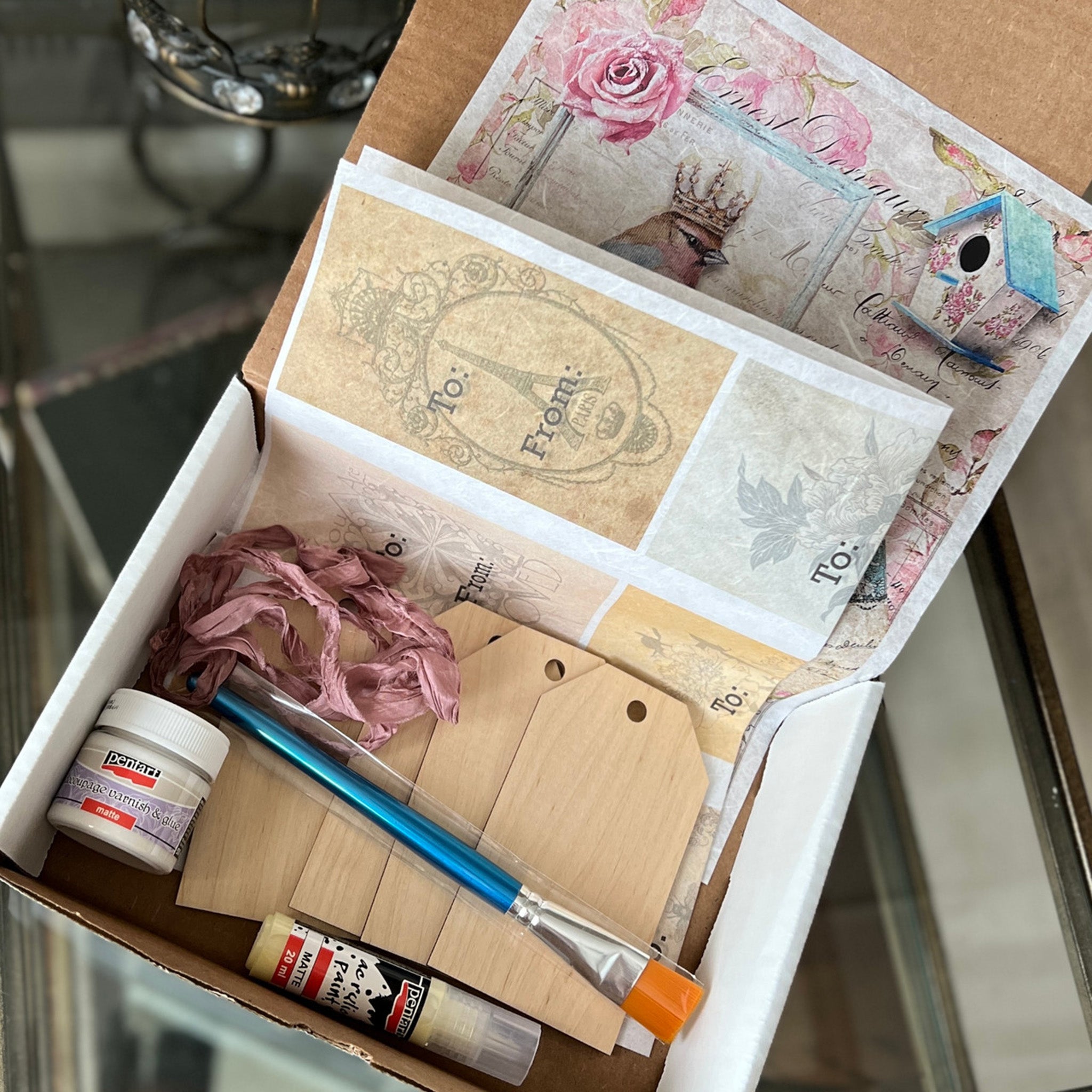A box of a decoupage beginner's kit that includes 4 wood gift tags, a sheet of To and From rice paper tags, decoupage glue, acrylic paint, a paintbrush, and an A4 rice paper featuring a French inspired design that includes pink roses, the Eiffel Tower, a birdhouse, and a bird wearing a crown.