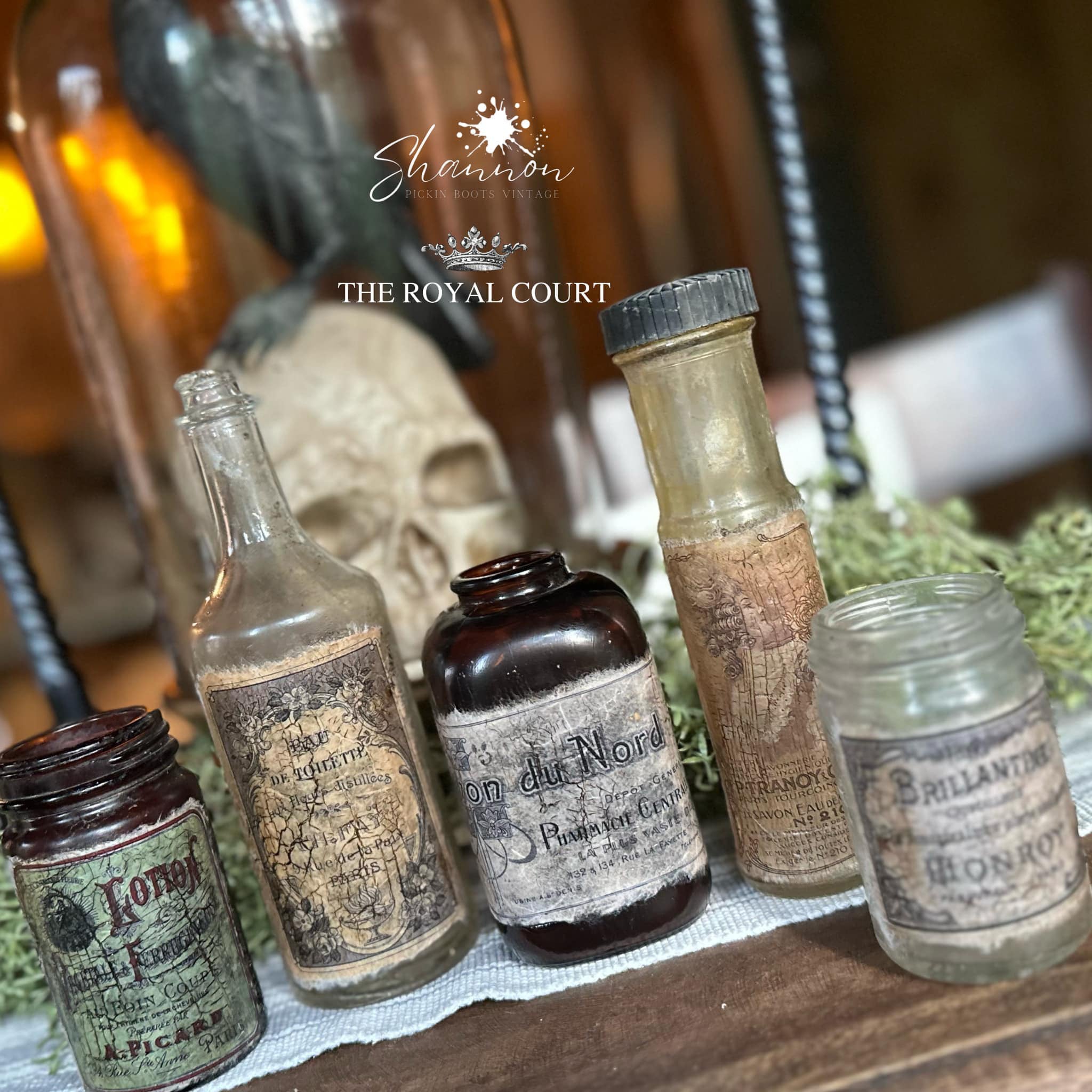 Five glass bottles and jars in front of a skull and crow decor piece feature Decoupage Queen's Perfume Labels rice paper on them.