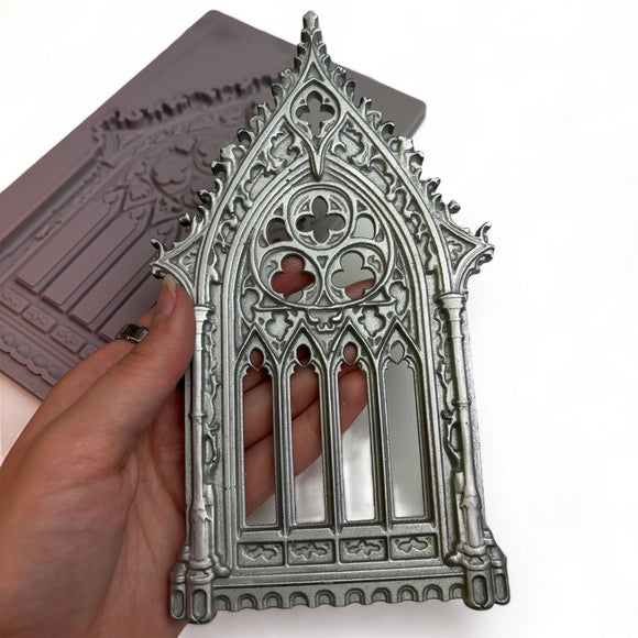 A hand is shown holding a silver colored silicone mold casting of an ornate Gothic window against a white background with the brown silicone mold.