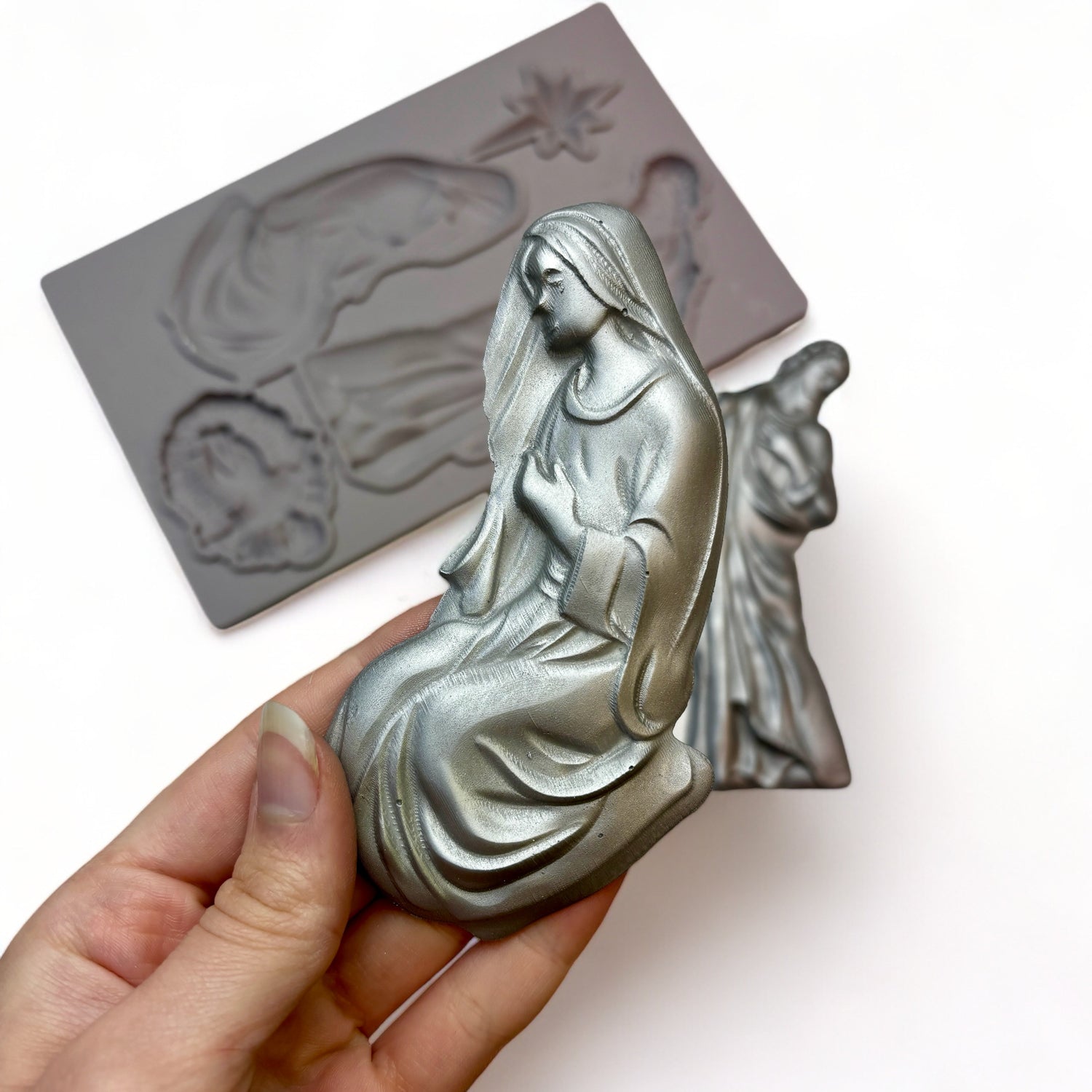 A beige silicone mold and silver colored castings of the Holy Family and the Bethlehem Star are against a white background. A hand is shown holding the casting of Mary.