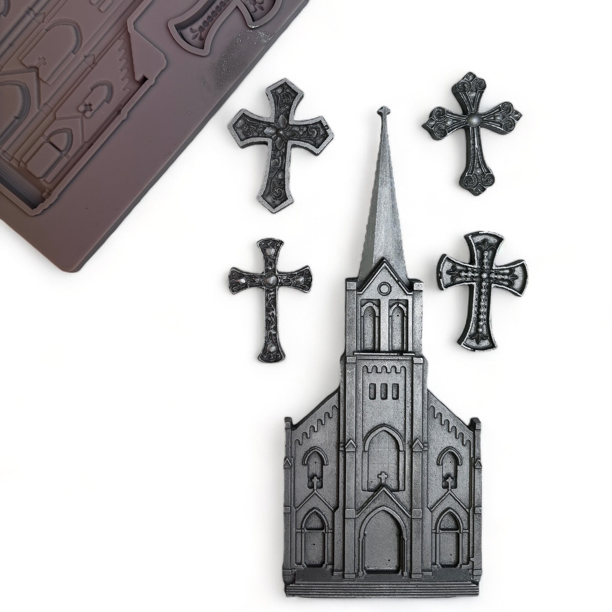 A brown silicone mold and silver colored castings of an old church with a tall steeple and 4 small ornate crosses are against a white background.