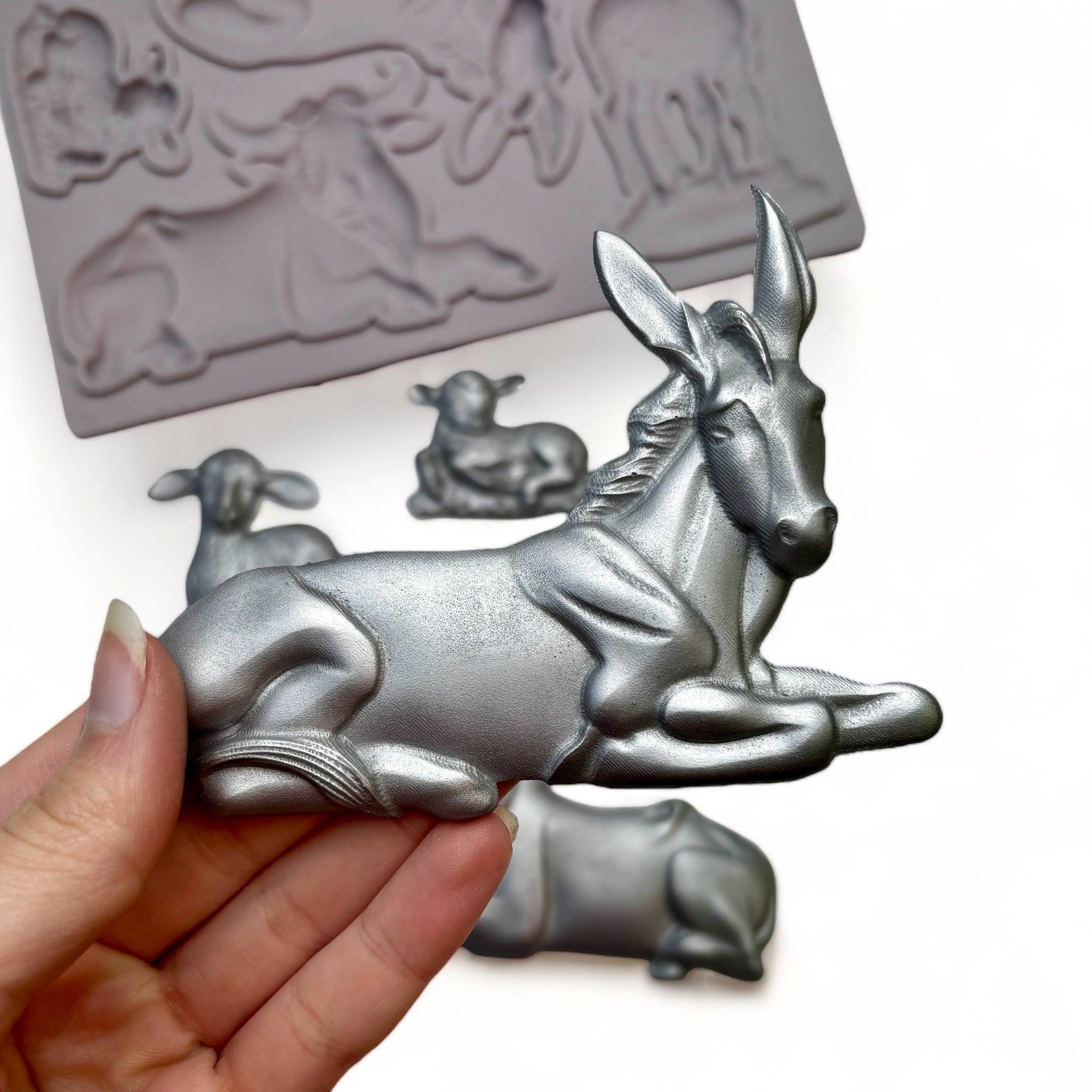 A beige silicone mold and silver colored castings of 4 nativity animals are against a white background. A hand is shown holding a donkey casting.