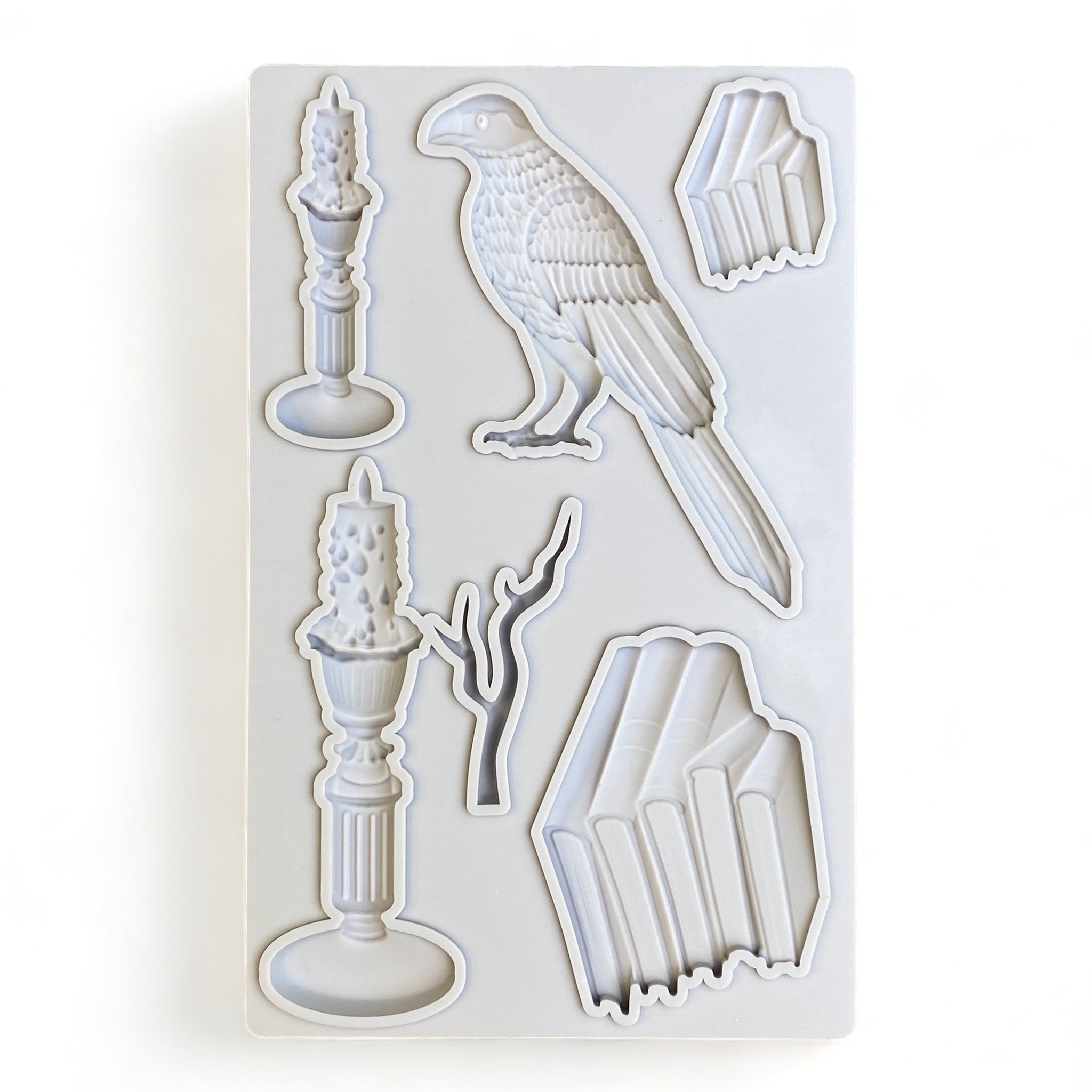 A light grey silicone mold of stacks of books, candles, a branch, and a bird is against a white background. 