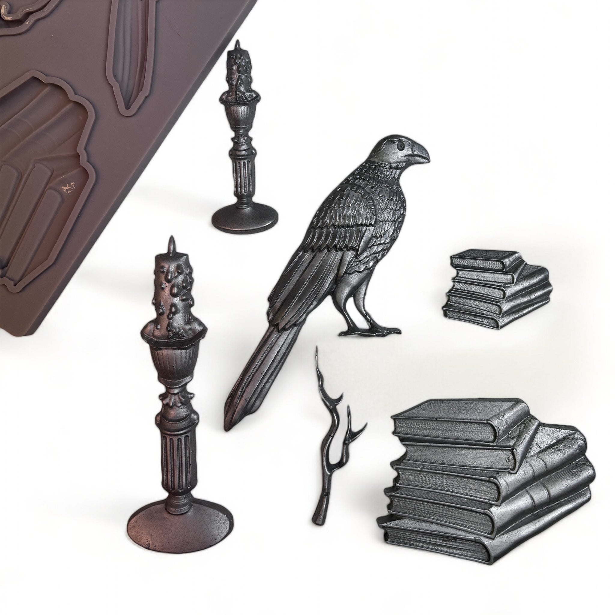 A brown silicone mold and silver colored castings of stacks of books, candles, a branch, and a bird are against a white background. 