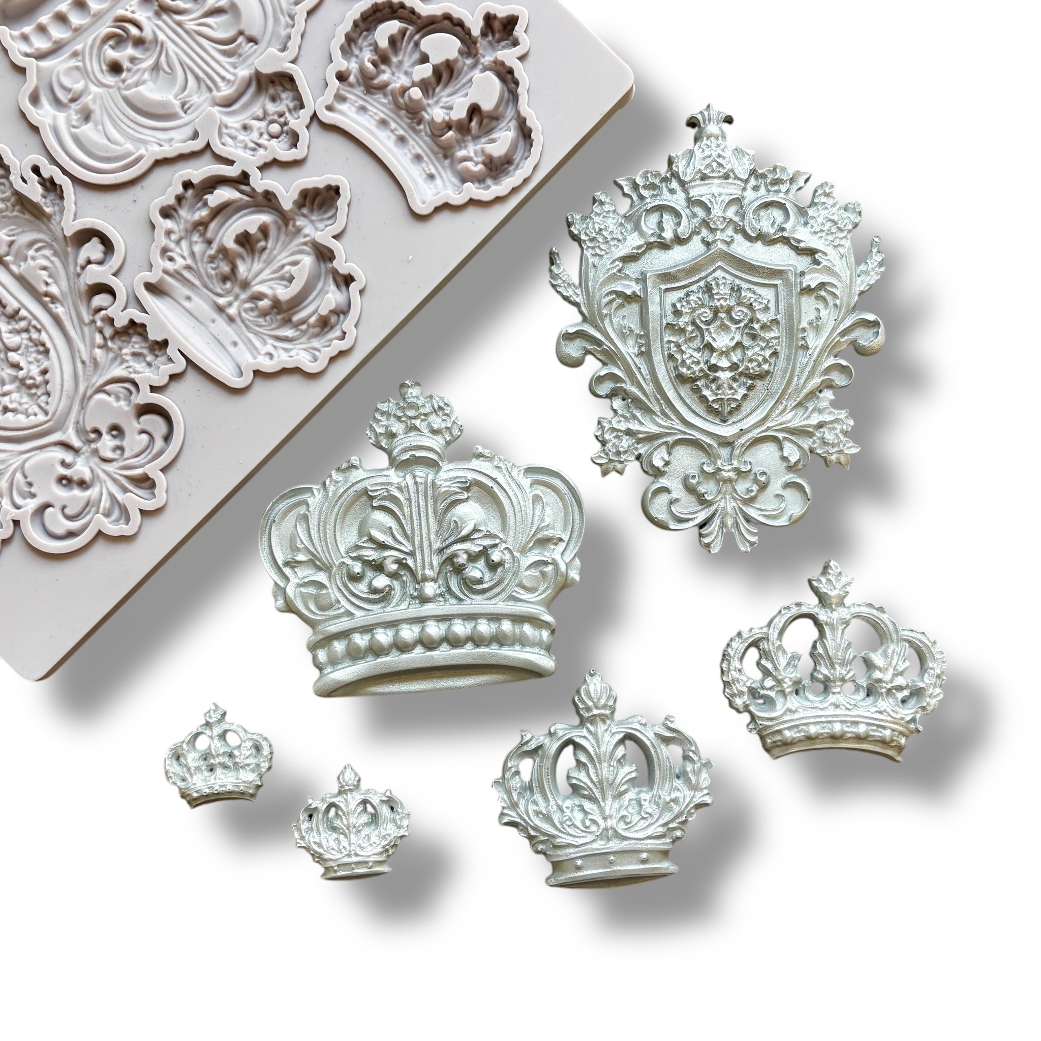 A light grey silicone mold and silver colored castings of 5 varying size regal crowns and a family crest are against a white background.