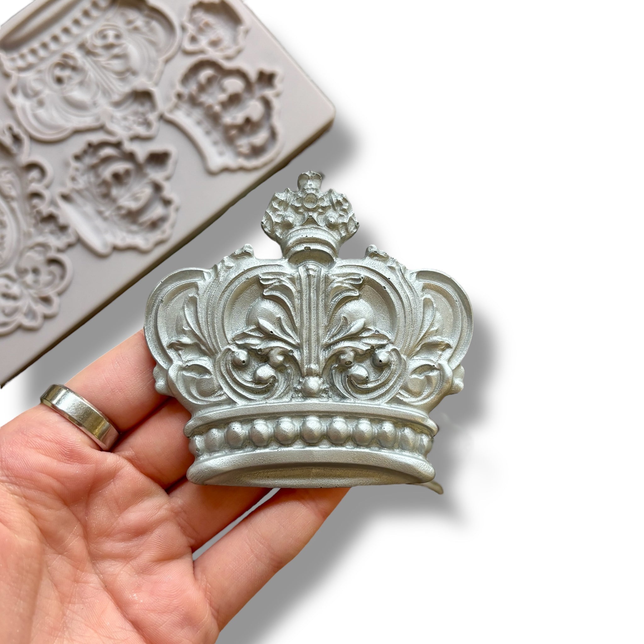 A light grey silicone mold featuring 5 varying size regal crowns and a family crest is against a white background. A hand is shown holding one of the silver colored crown castings.