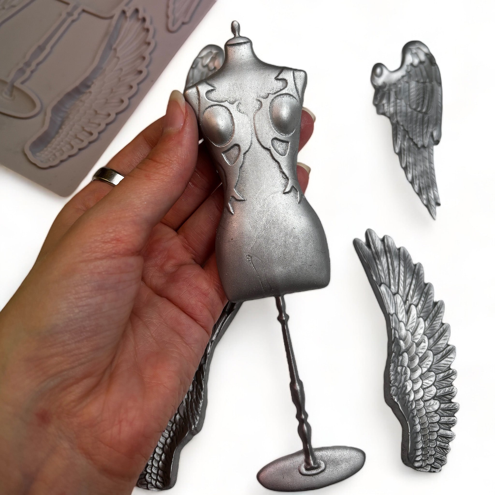 A brown silicone mold and silver colored castings of a seamstress dress form and 2 sets of angel wings are against a white background. A hand is shown holding the dress form