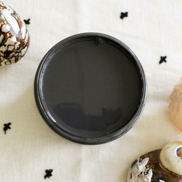 An arial view of an open container of Dixie Belle Paint Company's Black Sands All-In-One Silk Paint is on a white material with small back cross stitches. 