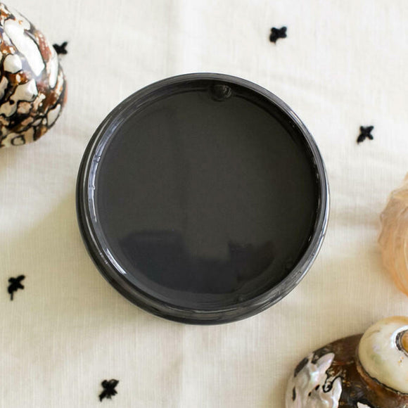 An arial view of an open container of Dixie Belle Paint Company's Black Sands All-In-One Silk Paint is on a white material with small back cross stitches. 