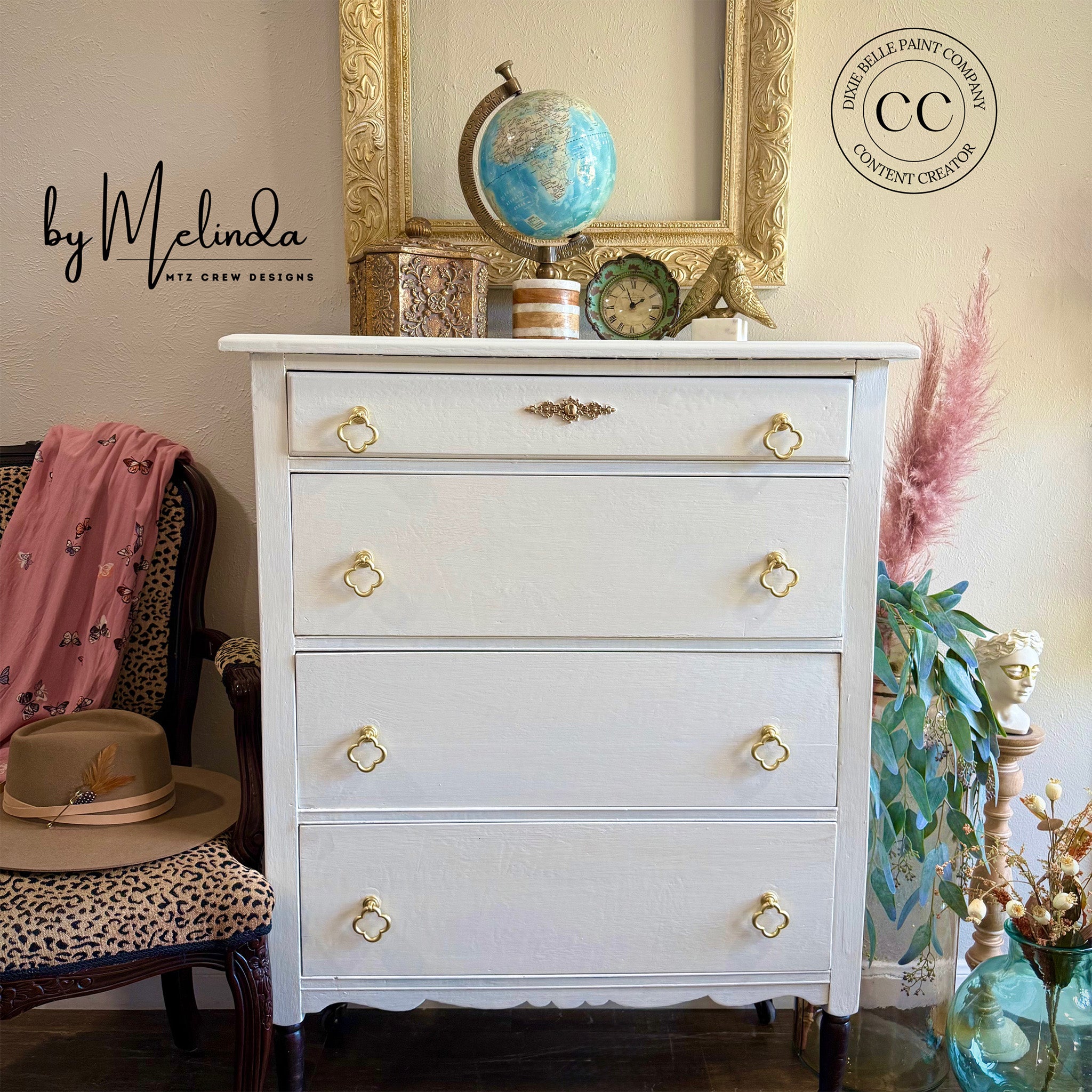 White dresser with gold knobs in a room with decor elements like a chair, plant, and framed picture.