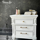 White dresser with decorative items against a dark wall with floral patterns, featuring the brand 'Theresa Lee'.