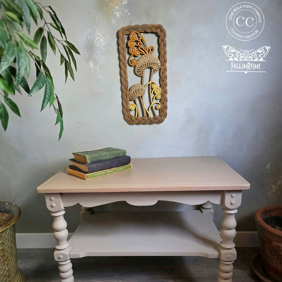 Decorative wall art above a table with books, surrounded by plants and a light gray wall.