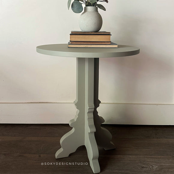 Round side table with a vase and books on a wooden floor against a white wall.
