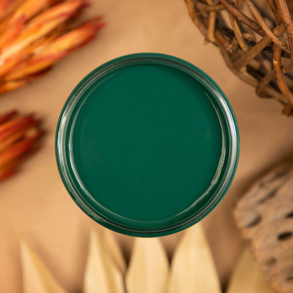 An arial view of an open container of Dixie Belle Paint Company's Midnight Green All-In-One Silk Paint is on a light brown table with natural wood and dried orange colored flowers.