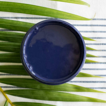 An arial view of an open container of Dixie Belle Paint Company's Nautical All-In-One Silk Paint is on a white material with dark grey stripes and a palm frond.