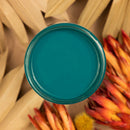An arial view of an open container of Dixie Belle Paint Company's Oasis All-In-One Silk Paint is on a sand brown table with dried palm fronts and orange colored dried flowers.