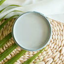 An arial view of an open container of Dixie Belle Paint Company's Nautical All-In-One Silk Paint is on a weaved wicker placemat with a white cloth napkin and green palm frond.