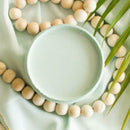 An arial view of an open container of Dixie Belle Paint Company's Tide Pool All-In-One Silk Paint is on a matching piece of material with a large light wood bead necklace and palm frond.