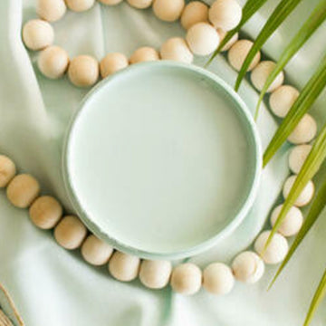 An arial view of an open container of Dixie Belle Paint Company's Tide Pool All-In-One Silk Paint is on a matching piece of material with a large light wood bead necklace and palm frond.