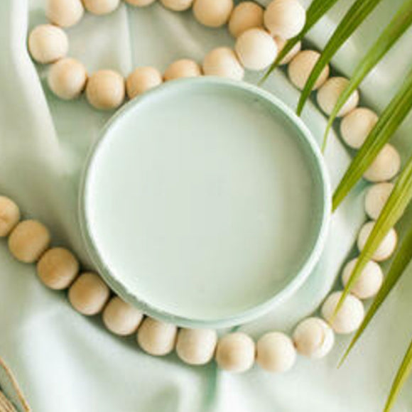 An arial view of an open container of Dixie Belle Paint Company's Tide Pool All-In-One Silk Paint is on a matching piece of material with a large light wood bead necklace and palm frond.
