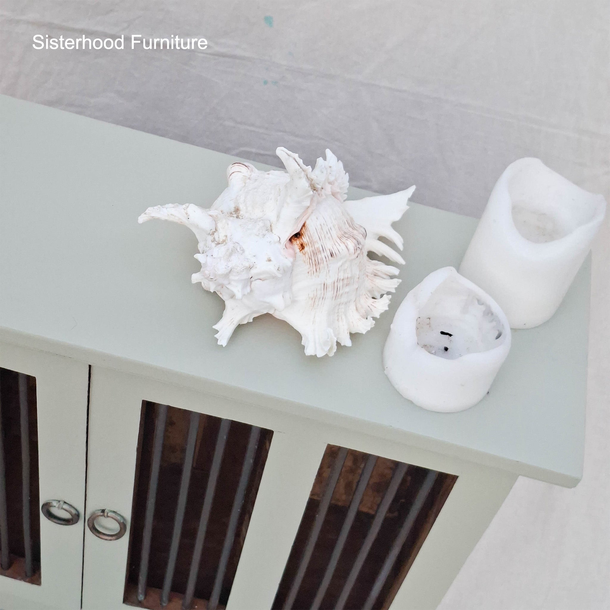 Decorative shell and candle holders on a light green surface with 'Sisterhood Furniture' branding.