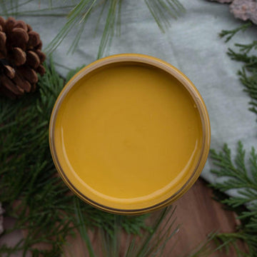 An arial view of an open container of Dixie Belle Paint Company's Yellowstone All-In-One Silk Paint is on a wood table with a grey cloth napkin, pine cone, and pine sprigs.