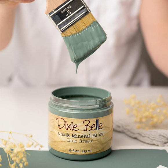 A paint brush with dripping paint is shown above an open container of Dixie Belle's Blue Grass Chalk Mineral Paint.