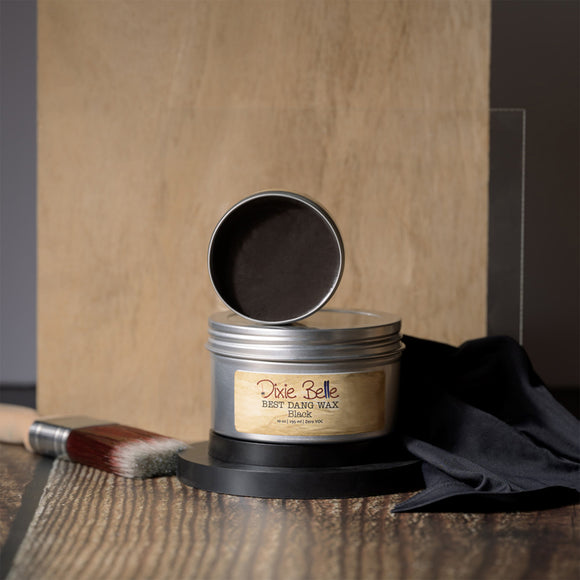 Metal tin with open lid containing a dark brown wax, branded 'Pixie Belle', on a wooden surface.