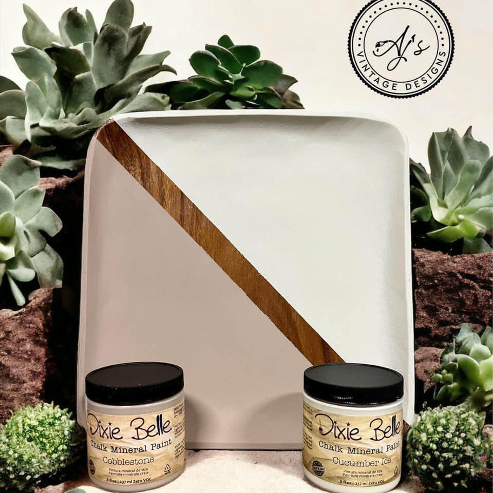 A square wood tray refurbished by Aj's Vintage Design is painted diagonally in Dixie Belle's Cobblestone chalk mineral paint on the bottom left, and Cucumber Ice chalk mineral paint on the top right with a natural wood stripe separating them. The tray is surrounded by green succulents.