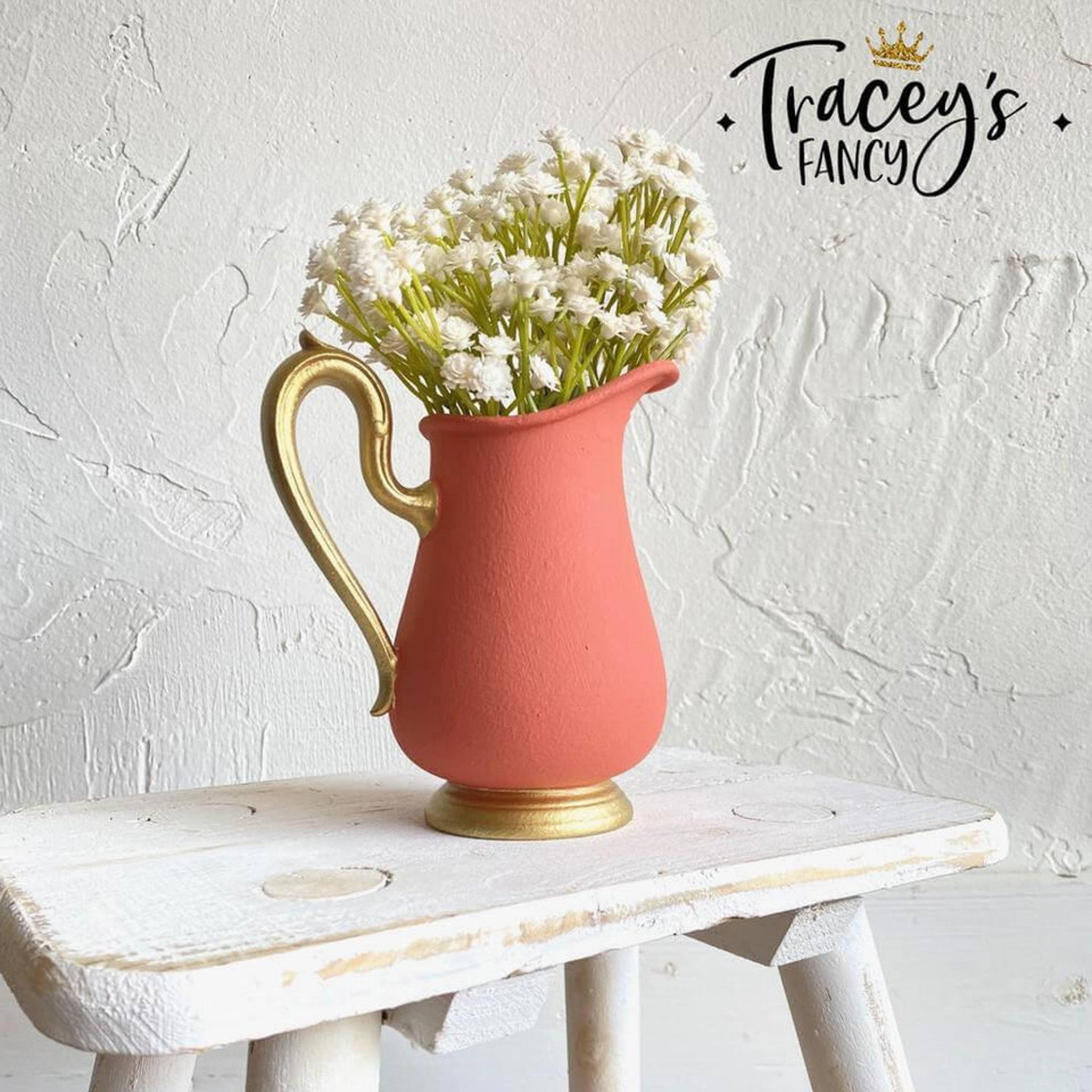 A ceramic water pitcher refurbished by Tracey's Fancy is painted with Dixie Belle's Cottage Door chalk mineral paint. The pitcher has a gold painted handle and base with white flowers coming out of the top.