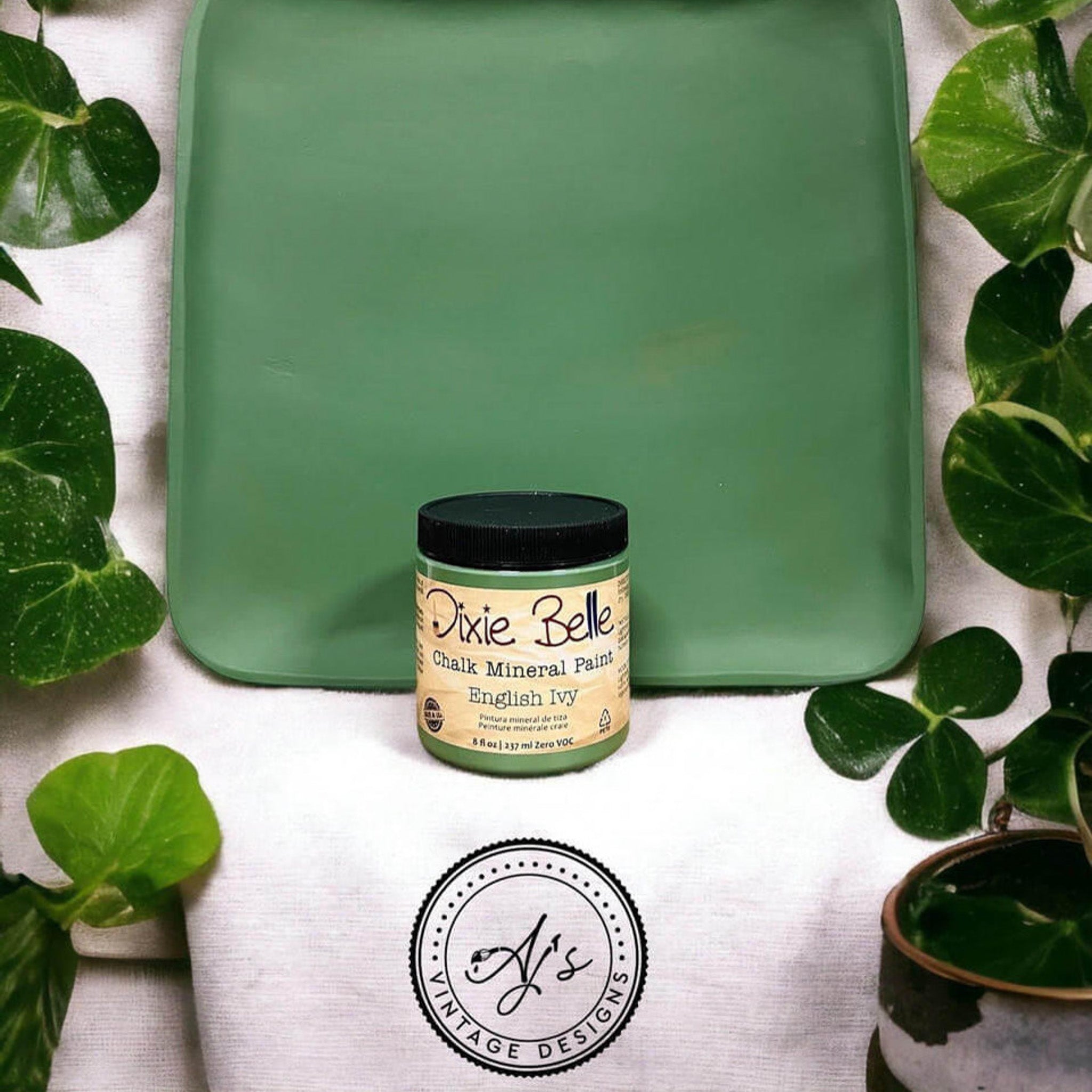 A wood tray repainted by AJ's Vintage Designs using Dixie Belle's English Ivy chalk mineral paint is pictured with a container of the paint. Green foliage surround the tray.