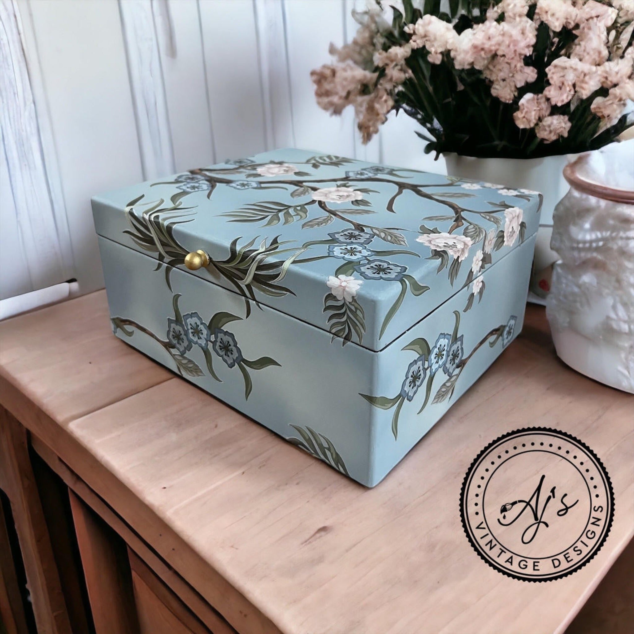 A wood box on a table refurbished by AJ's Vintage Designs is painted light blue and features Belles & Whistles' Chinoiserie rub-on transfer on it.