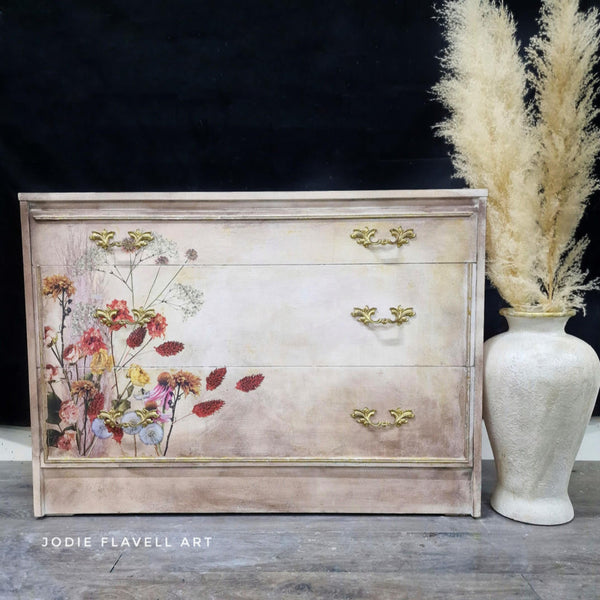 A 3-drawer dresser defurbished by Jodie Flavell Art is painted a blend of beige and white and features the Pink Pampas and Dried Wilds on its drawers.