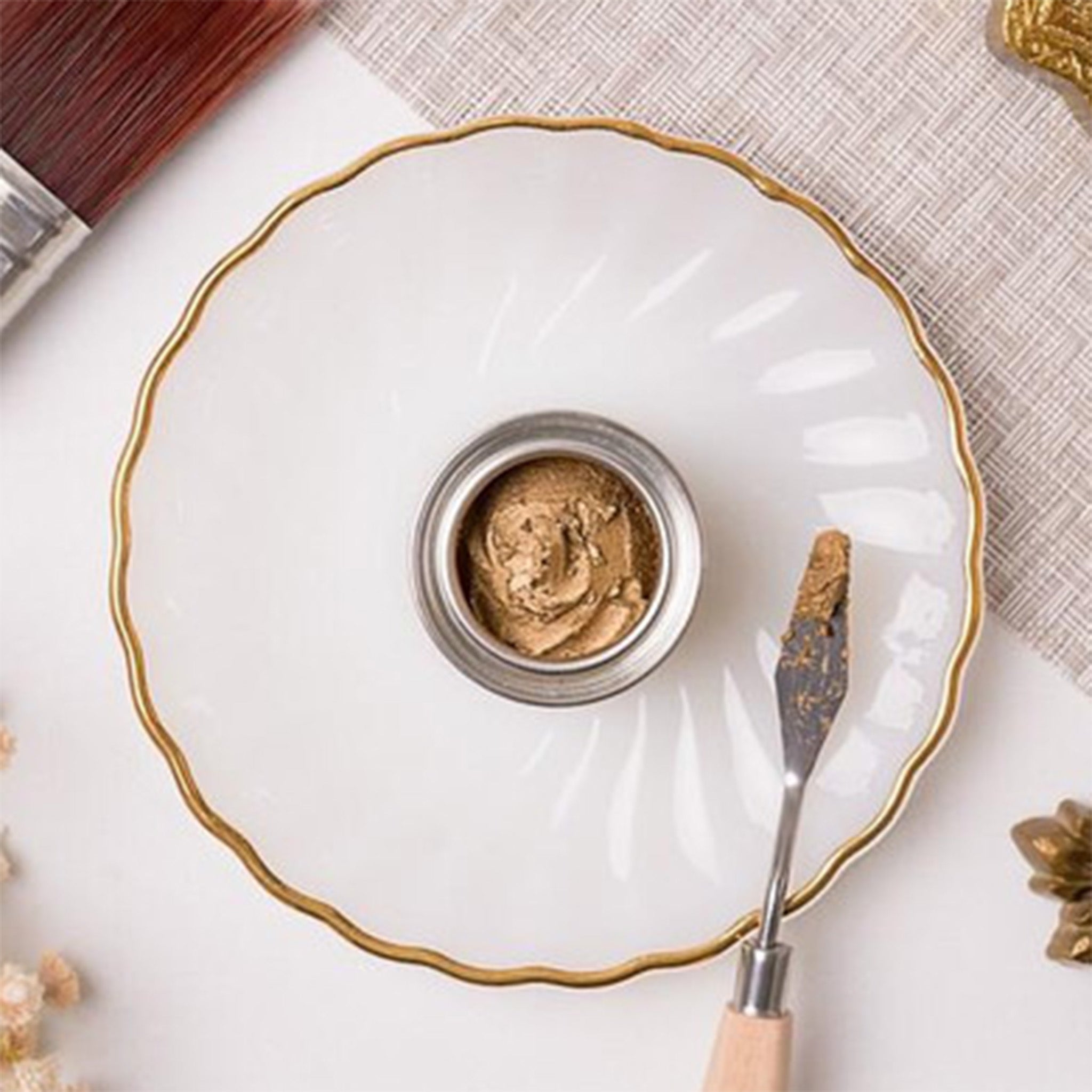 An open can of Dixie Belle's Antique Gold Gilding Wax is sitting on a white scalloped plate with a sample of the gilding wax on the edges. A pallet knife is shown with the wax on it.