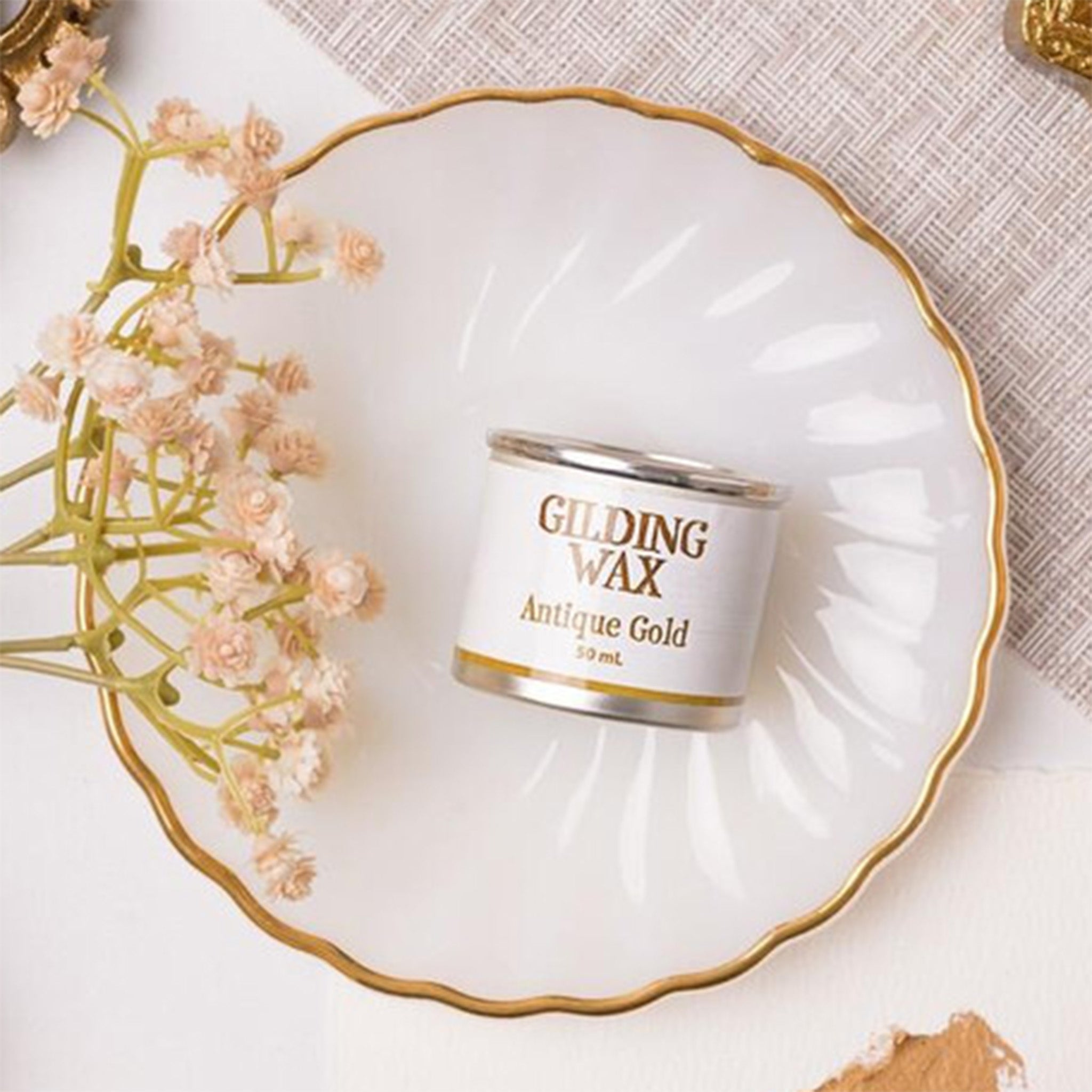 A can of Dixie Belle's Antique Gold Gilding Wax is laying on a white scalloped plate with a sample of the gilding wax on the edges.