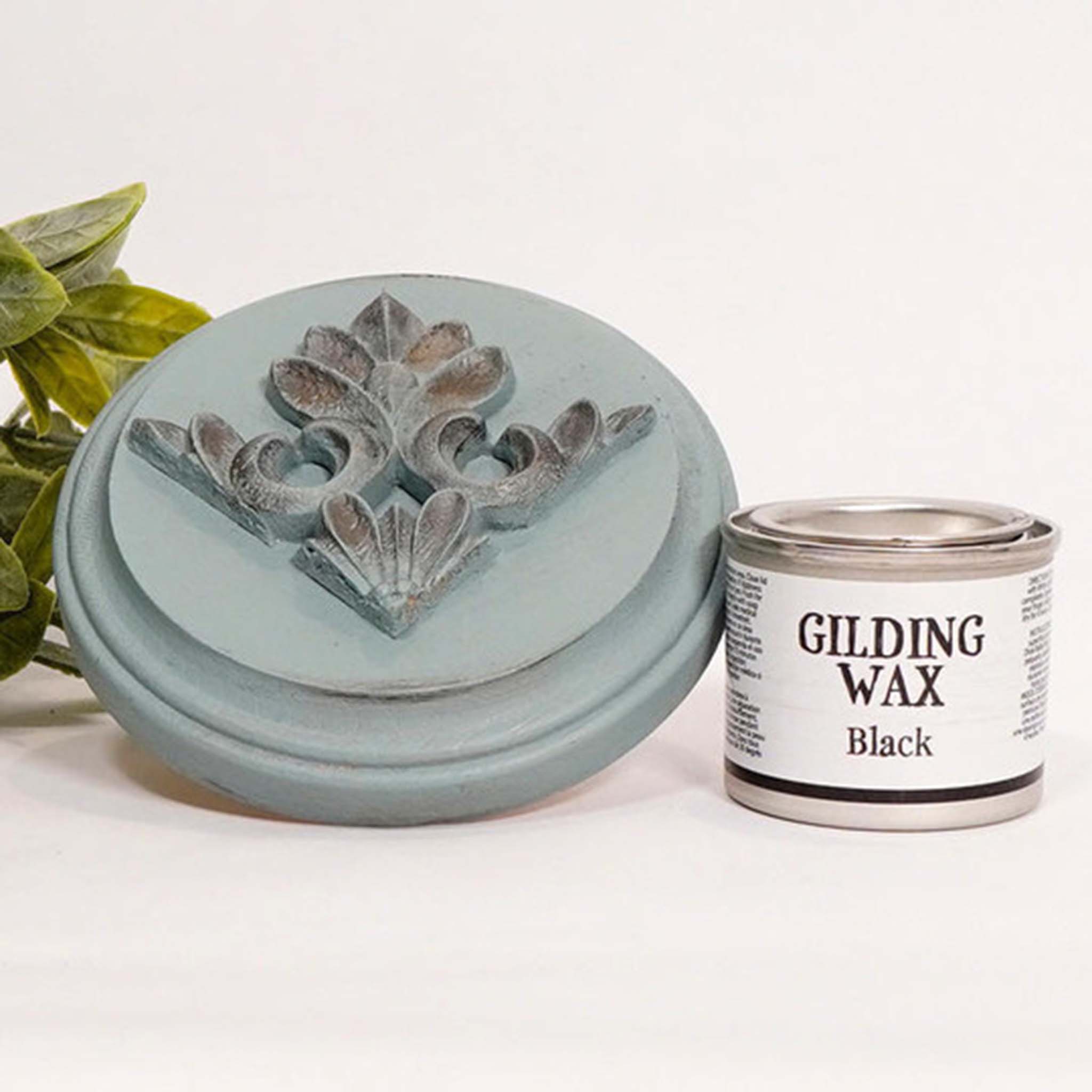 A can of Dixie Belle's Black Gilding wax is next to a light blue painted round wood sample. The wood sample has an ornate silicone mold casting that features the black gilding wax on it.