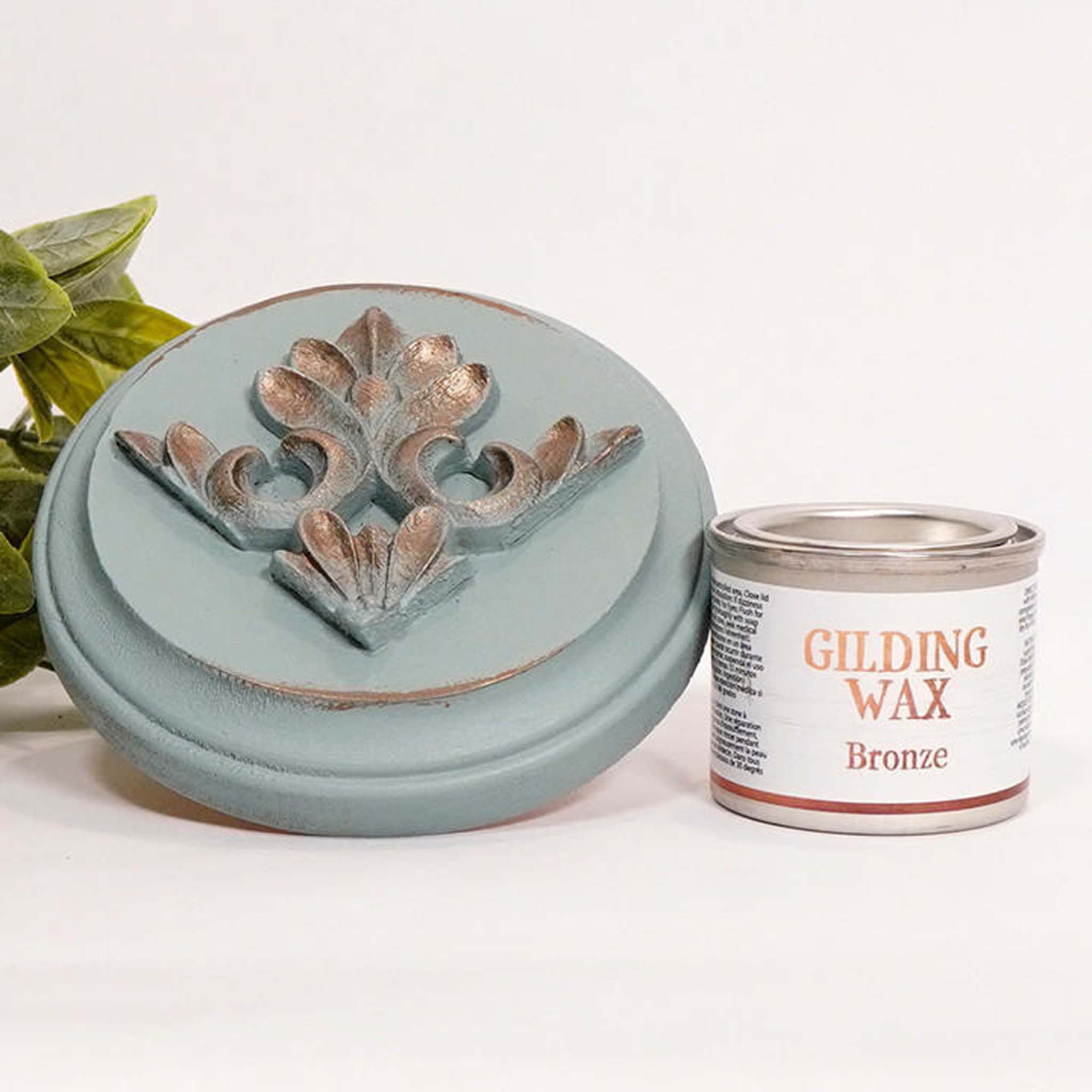 A can of Dixie Belle's Bronze Gilding wax is next to a light blue painted round wood sample. The wood sample has an ornate silicone mold casting that features the bronze gilding wax on it.