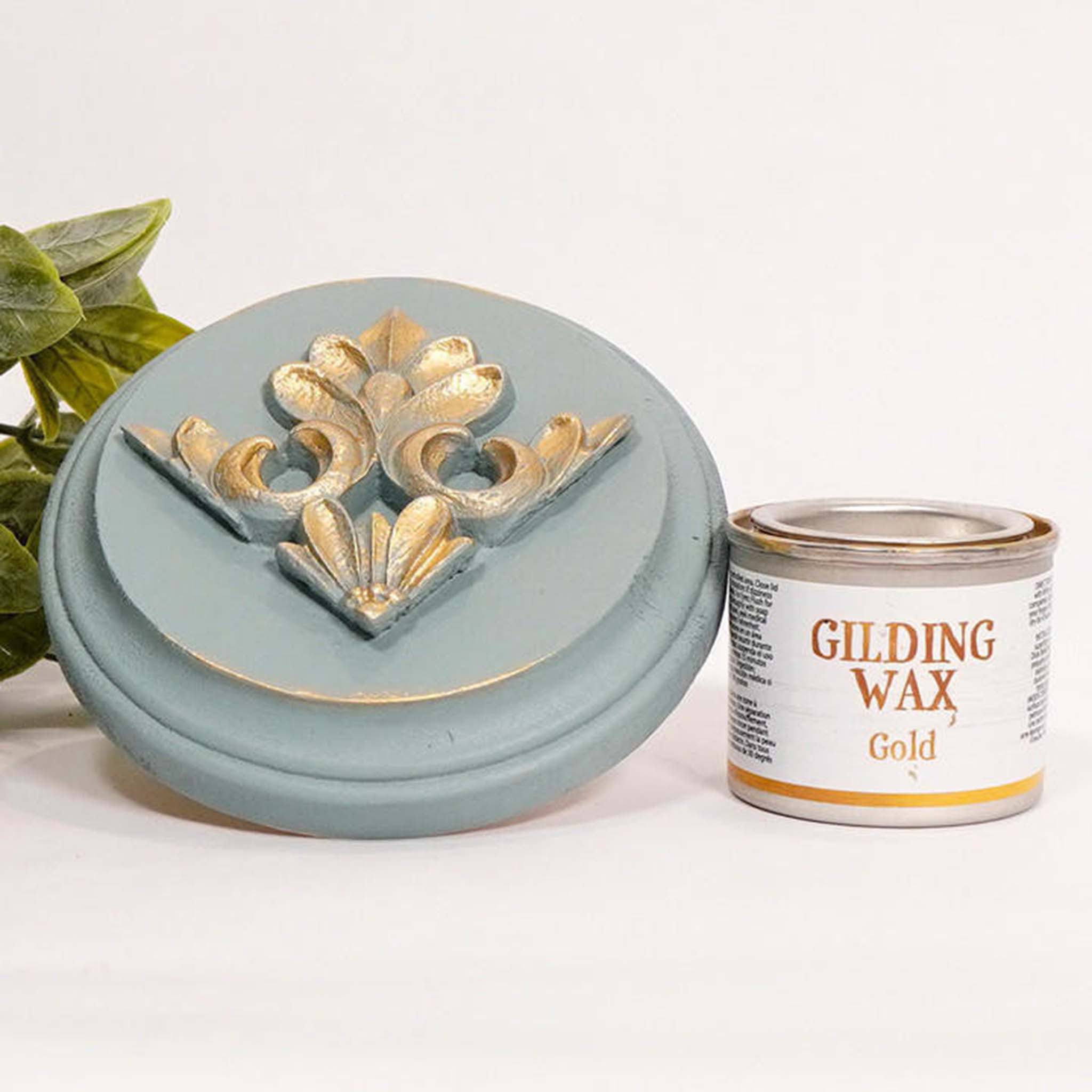 A can of Dixie Belle's Gold Gilding wax is next to a light blue painted round wood sample. The wood sample has an ornate silicone mold casting that features the gold gilding wax on it.