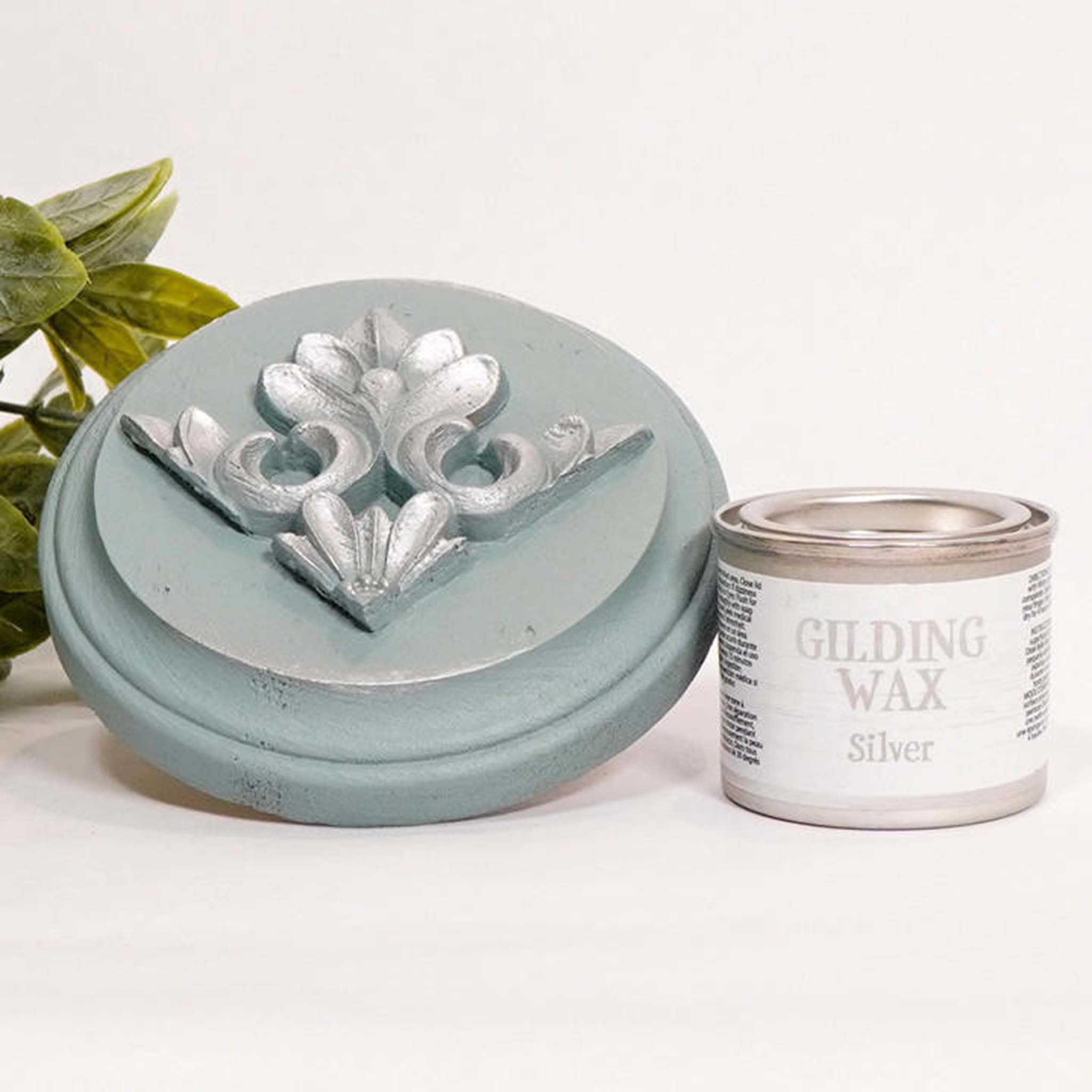 A can of Dixie Belle's Silver Gilding wax is next to a light blue painted round wood sample. The wood sample has an ornate silicone mold casting that features the silver gilding wax on it.