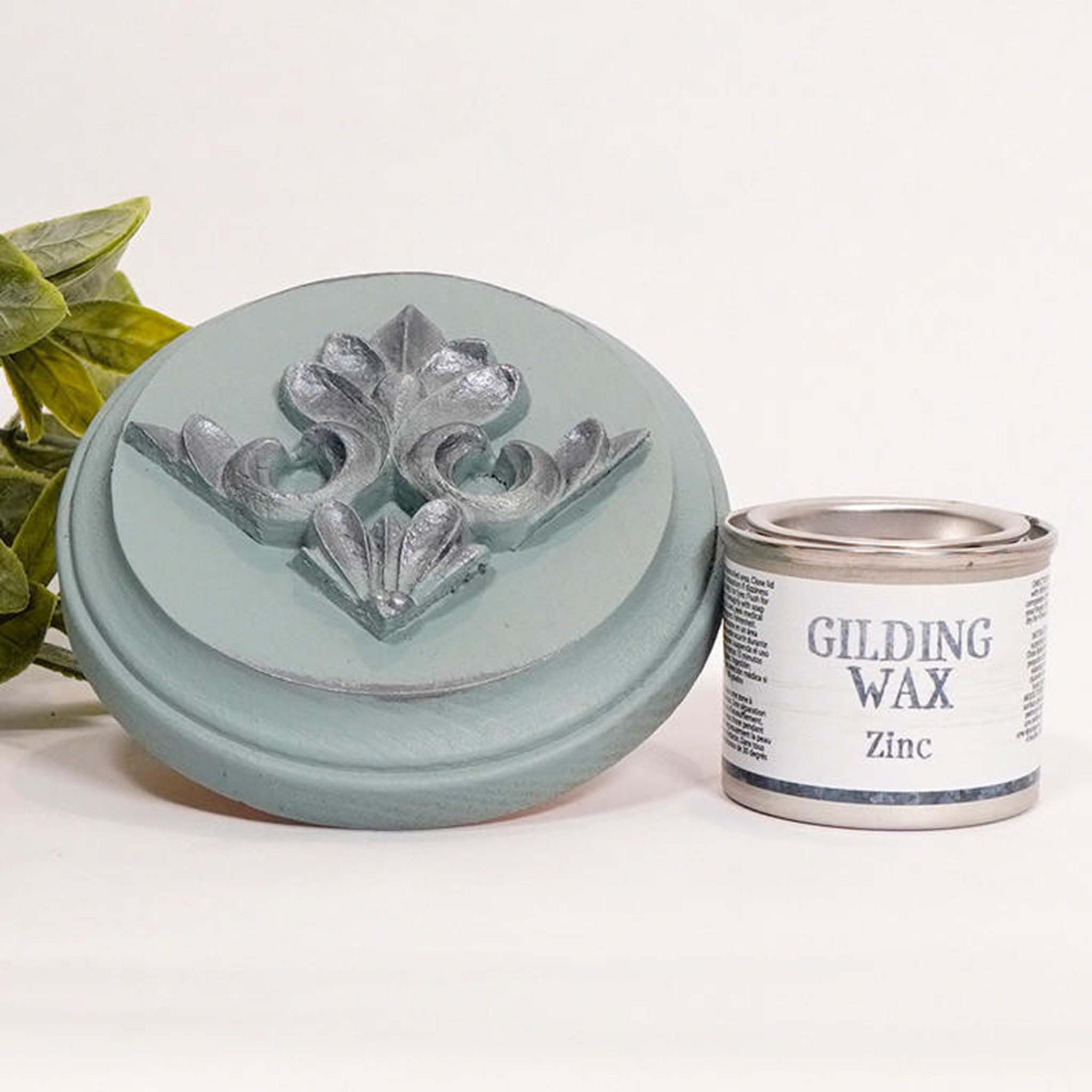A can of Dixie Belle's Zinc Gilding wax is next to a light blue painted round wood sample. The wood sample has an ornate silicone mold casting that features the zinc gilding wax on it.
