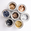 Open jars of Dixie Belle Paint Gilding Wax in metallic colors (gold, copper, bronze, silver, zinc, black) on a white background