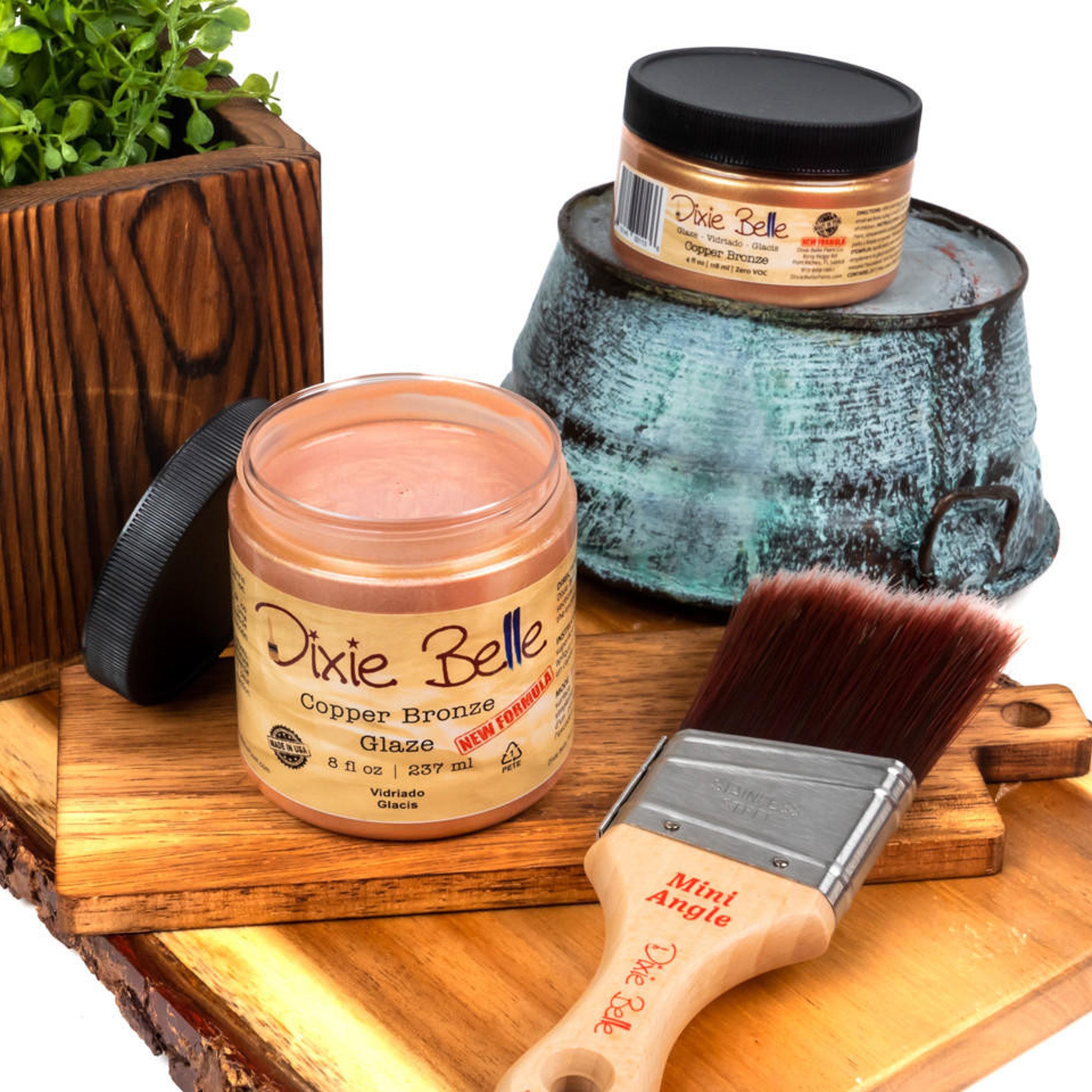 An 8oz/237ml and 4oz/118ml of Dixie Belle Paint's Copper Bronze Glaze are featured on a wood shelf with a Dixie Belle mini Angle paint brush.