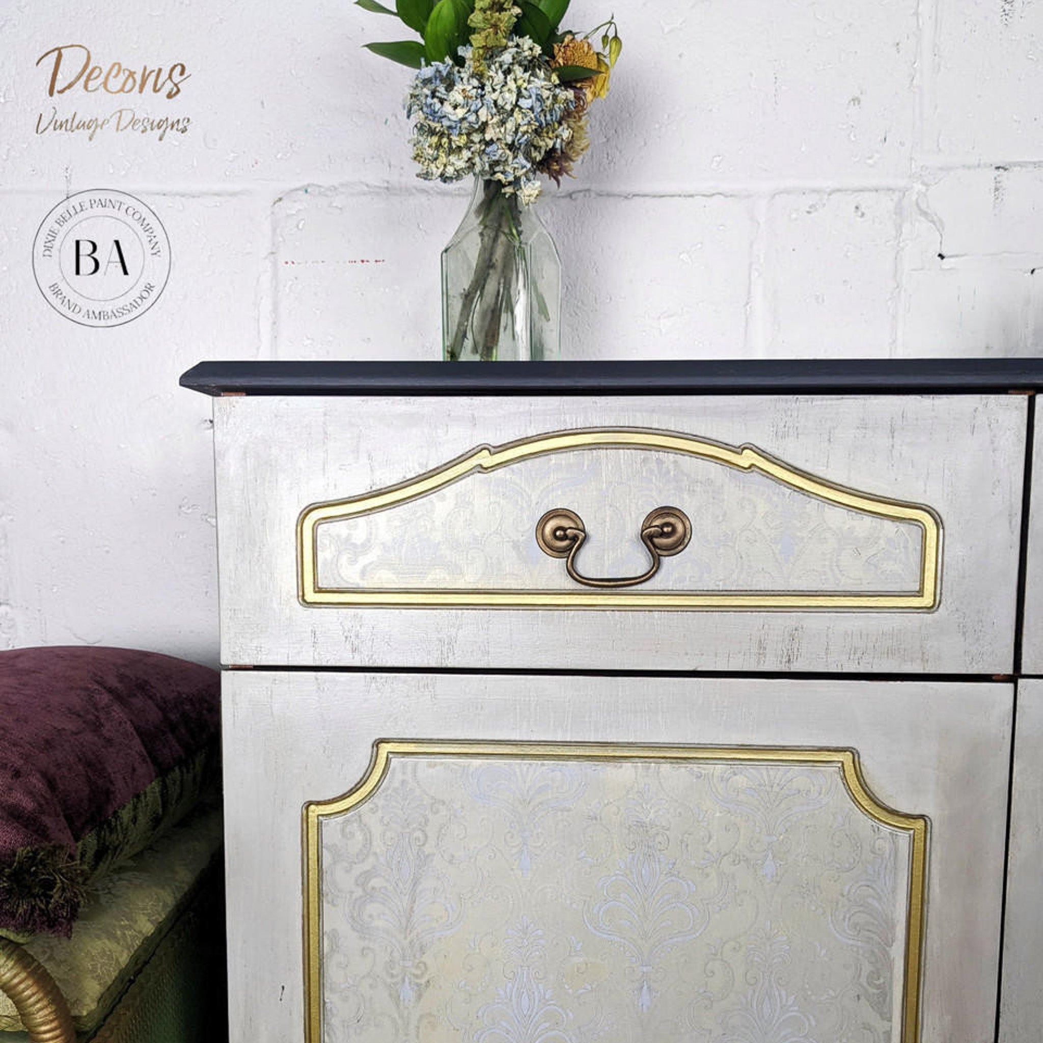 A close-up of a vintage dresser refurbished by Decoris Vintage Designs is painted light grey with gold accents and features Dixie Belle Paint's Hi Ho Silver Glaze as a stencil design on the door and drawer center inlays.
