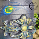 A close-up view of a vase refurbished by The Purple Posie has a large silicone mould casting on it and the entire piece is covered in Dixie Belle Paint's Hi Ho Silver Glaze. The mould has yellow and blue accents on it.