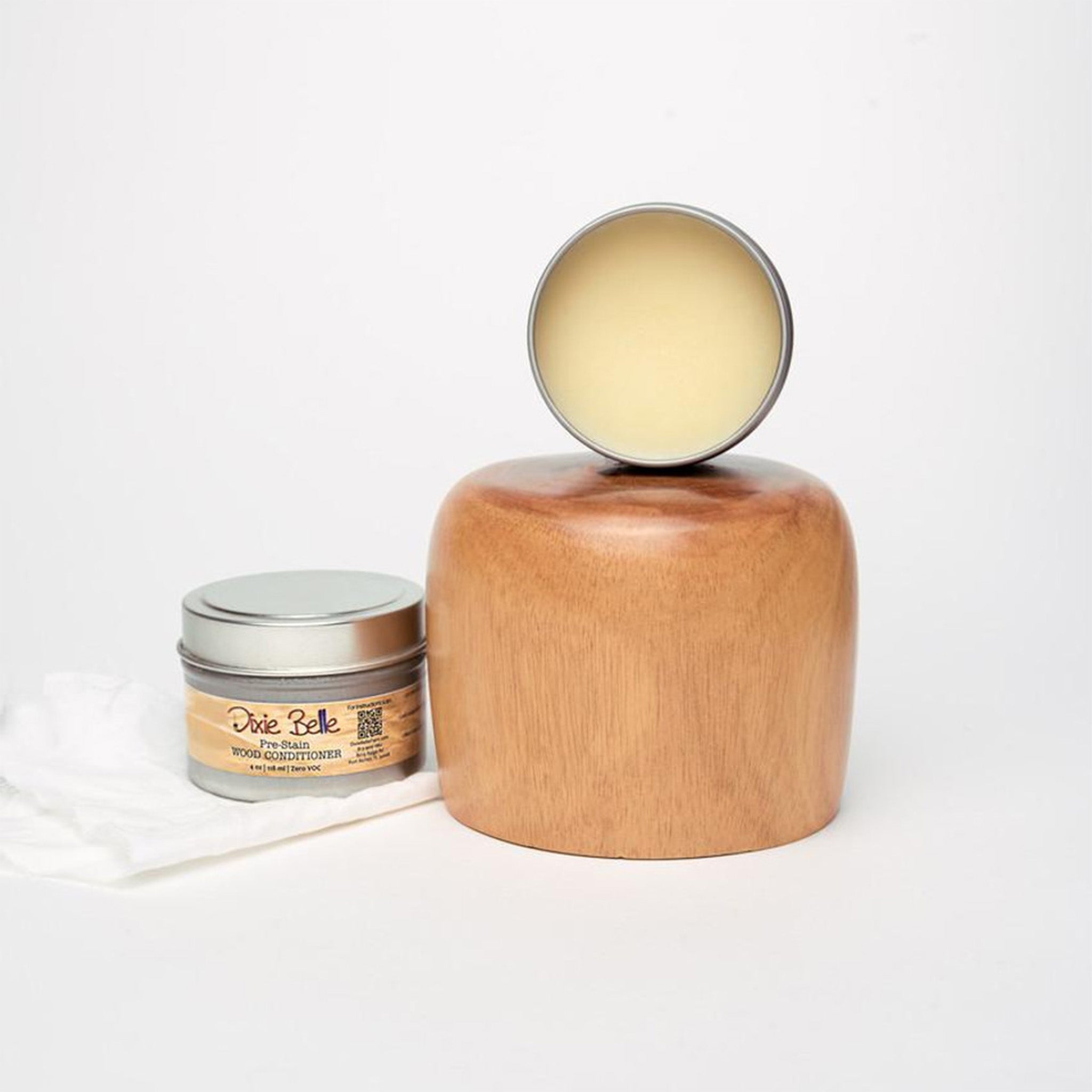 Two containers of Dixie Belle Paint's Pre-Stain Wood conditioner are against a white background. A closed container sits on a paper towel and an open container sits on a rounded wood block.