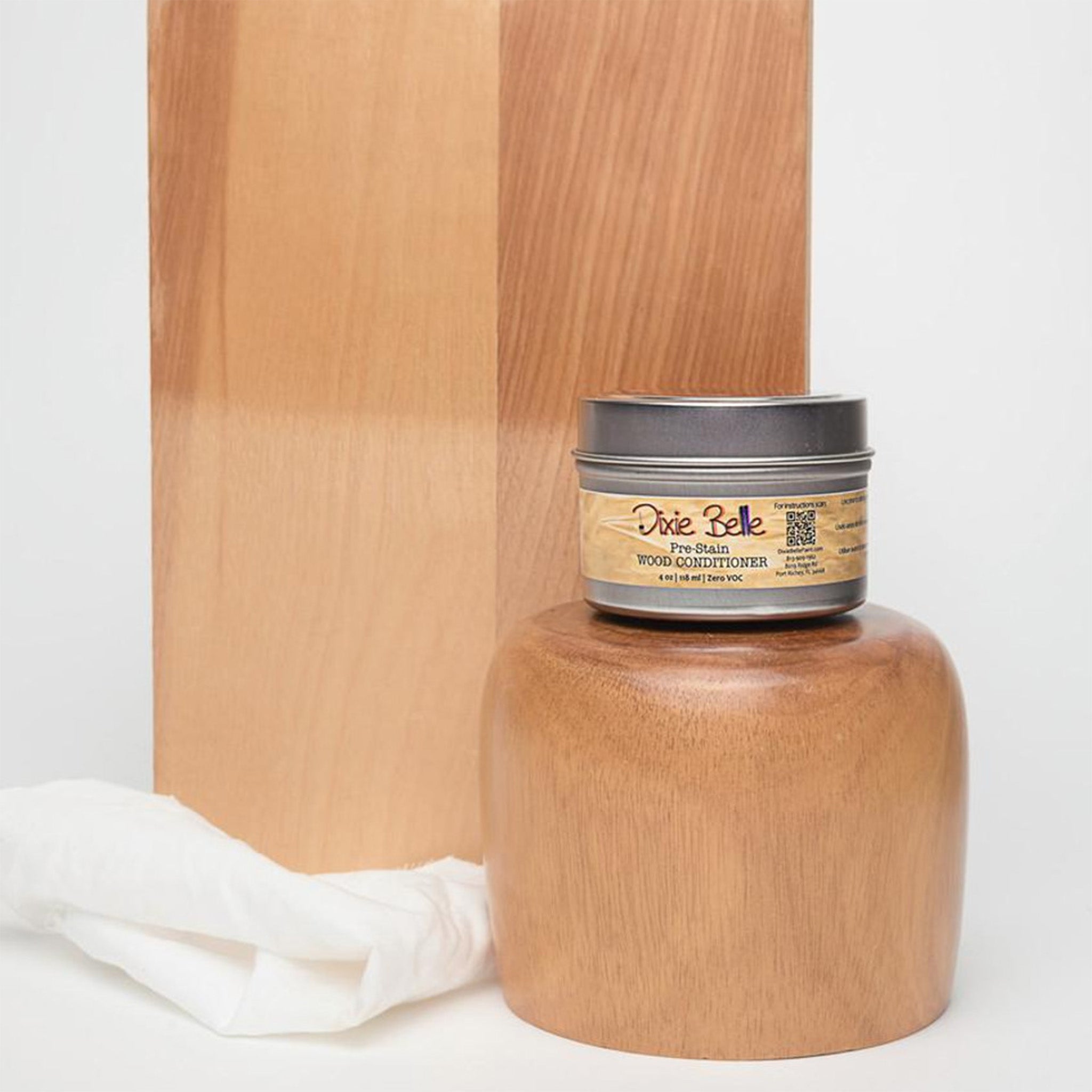 A 4oz container of Dixie Belle Paint's Pre-Stain Wood Conditioner is sitting on a cylindrical wood piece against a white background. Behind it is a sample of wood showing the top half conditioned using the product. 